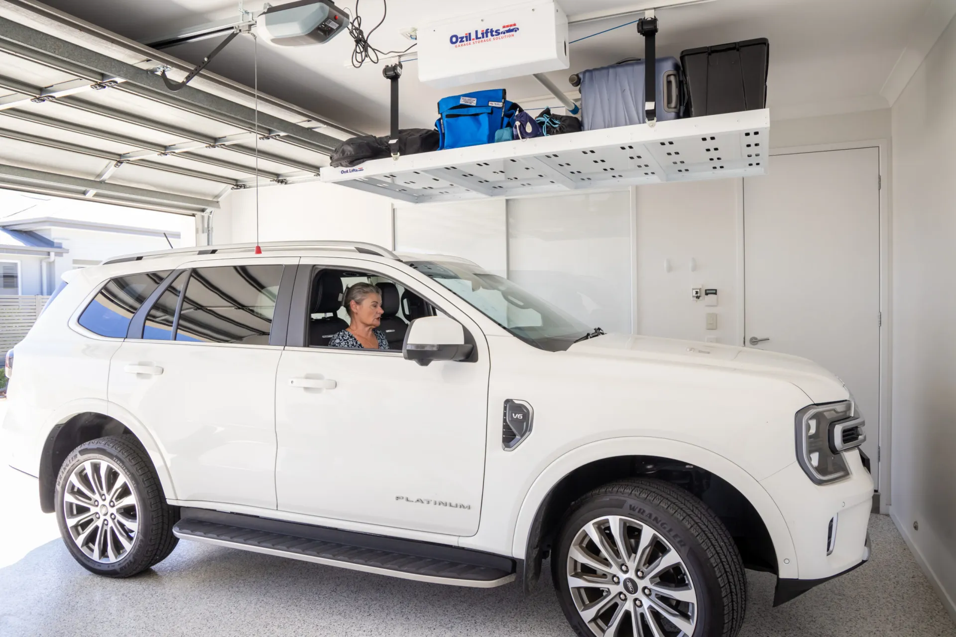 White Ford SUV parked in a bright garage, a woman sits in the driver’s seat beneath an overhead storage platform with bags and a white box labeled Ozil Lifts