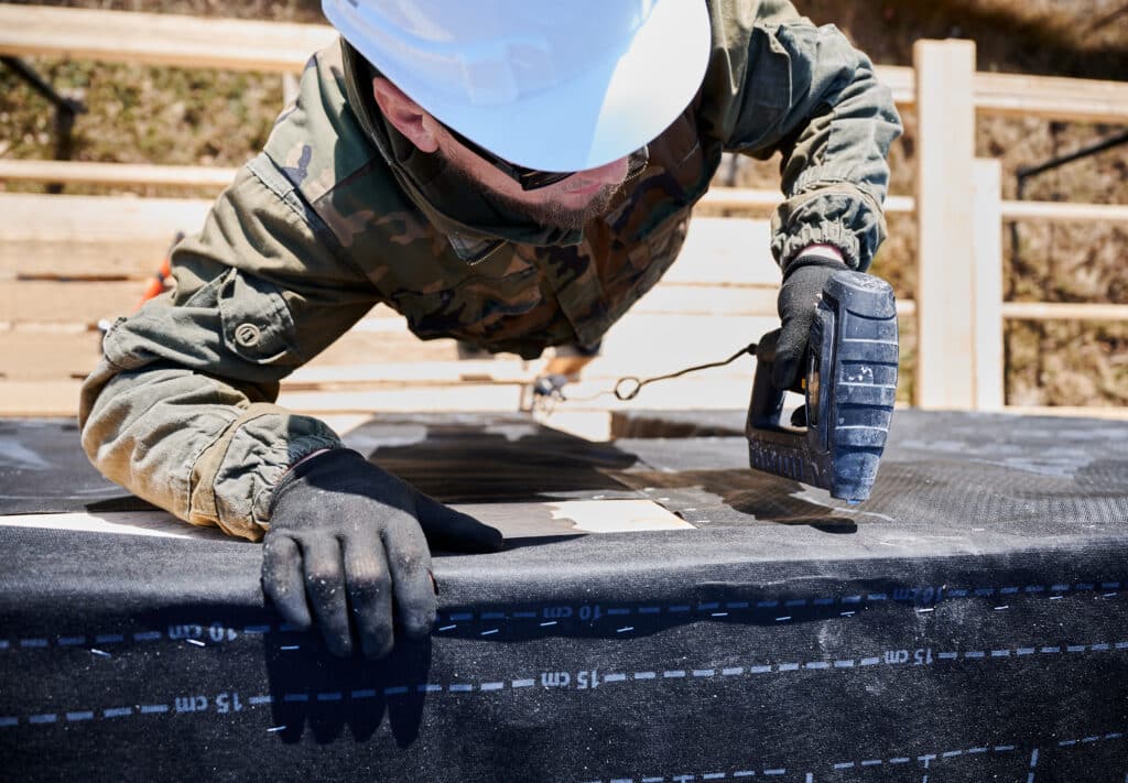 Male builder installing waterproof membrane on wall of future wooden frame house.