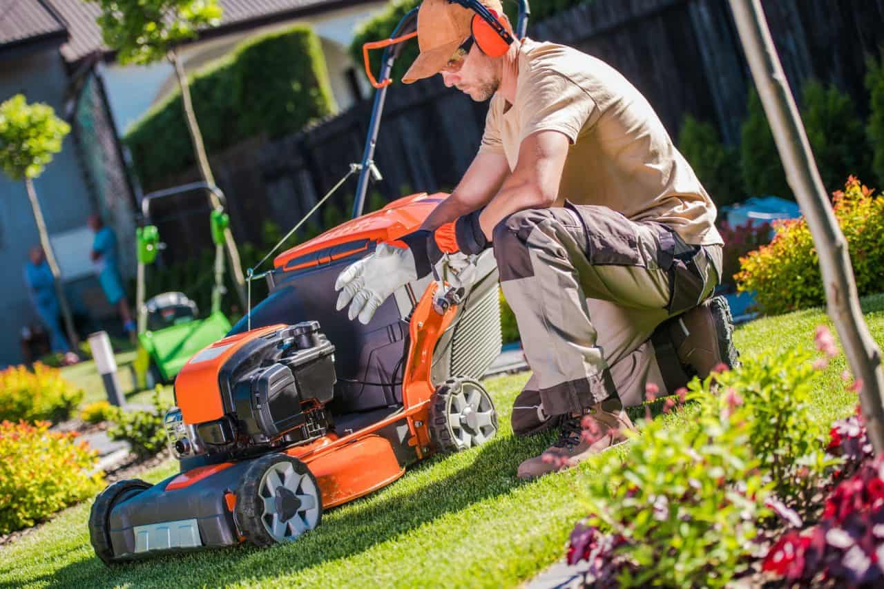 Une personne portant des protections auditives et des gants s'accroupit à côté d'une tondeuse à gazon orange sur une pelouse bien tondue, inspectant ou réglant la machine. Jardin avec diverses plantes vertes et buissons en arrière-plan.