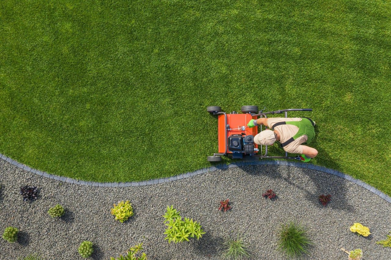 Une personne portant un chapeau et une combinaison verte tond une pelouse bien entretenue avec une tondeuse à gazon orange près d'une bordure de gravier gris et de petites plantes.