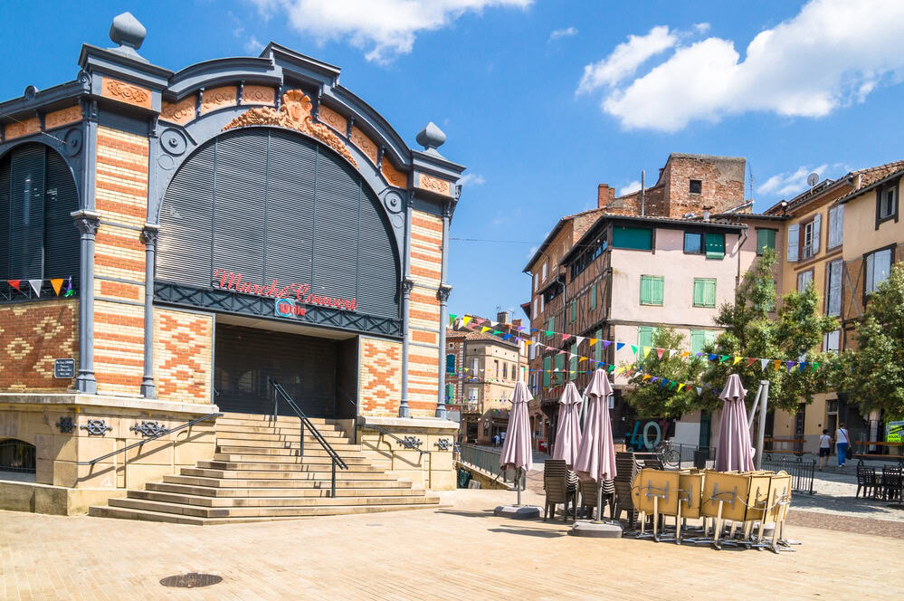 Une petite place vide avec un vieux bâtiment avec des marches en arrière-plan, des tables pliantes et des parasols fermés, des drapeaux colorés et des bâtiments rustiques sous un ciel ensoleillé avec des nuages.