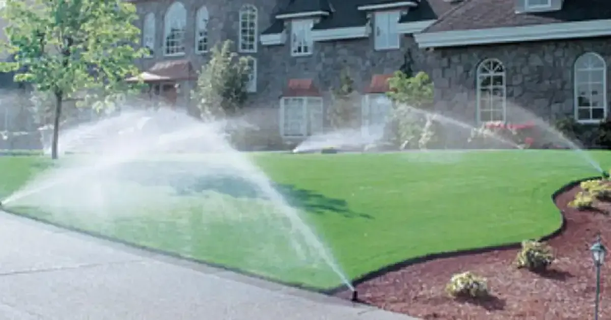 Sprinklers watering a well-manicured lawn in front of a large stone house with multiple windows and a steep roof, perfect for homeowners who value their garden's beauty. Remember, Sprinkler System Winterization in West Bloomfield is crucial to maintaining this pristine lawn year-round.