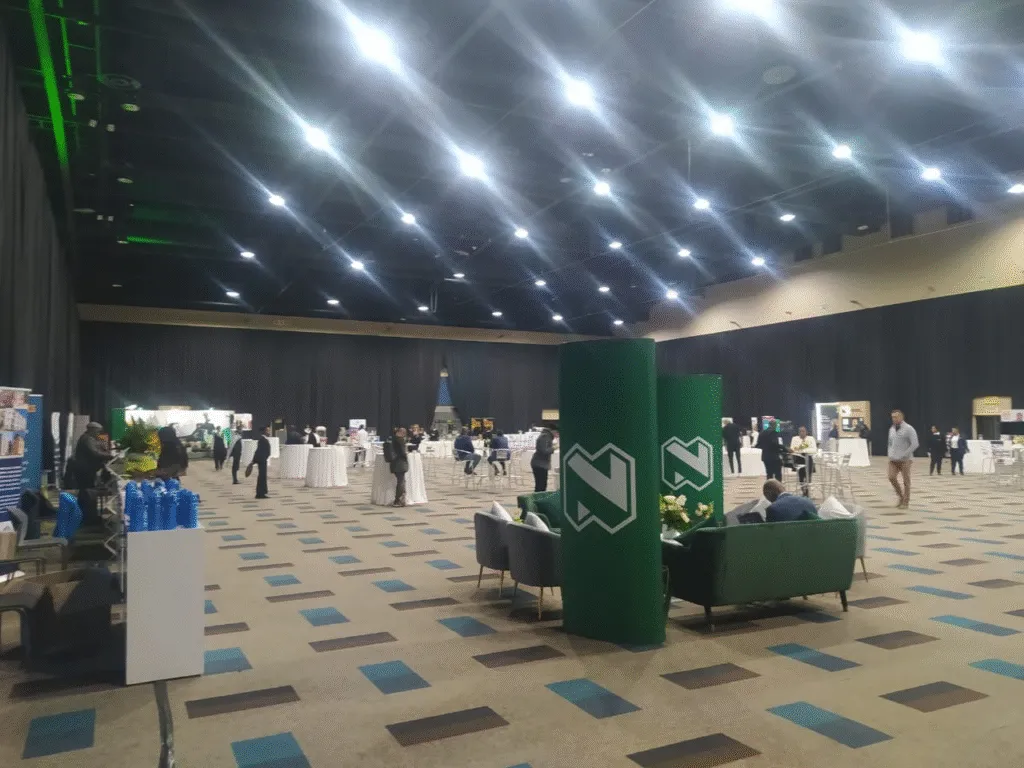 Large indoor event space with exhibition stands, scattered attendees, lounge seating, and branded green column displays under bright ceiling lights. - Proudly Developing