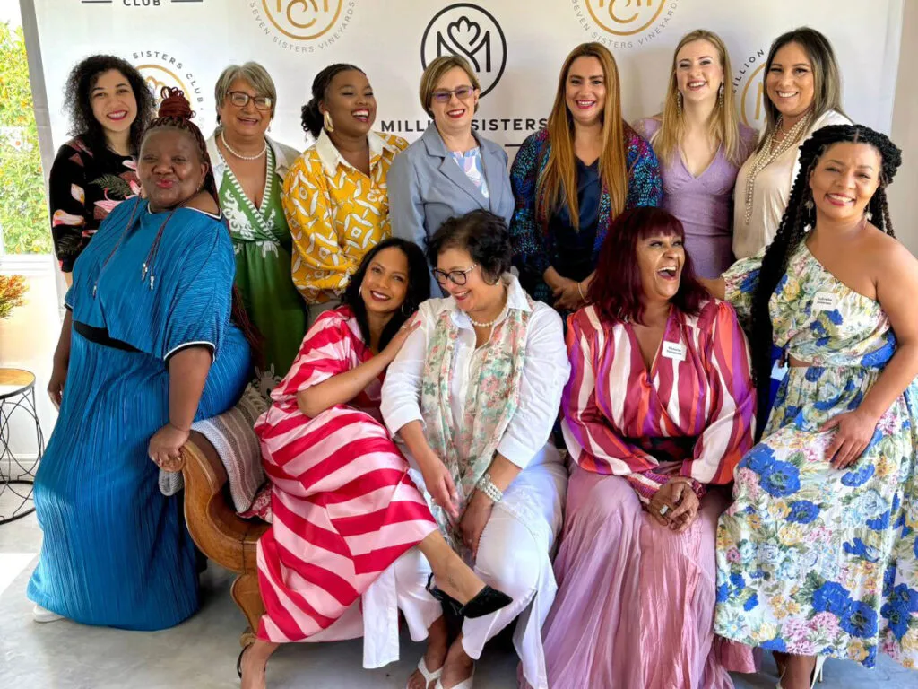 Thirteen women in colourful dresses pose and smile together indoors in front of a backdrop at a group event, celebrating the spirit of women’s empowerment events. - Proudly Developing