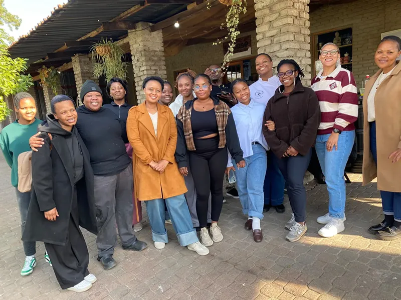 A group of thirteen people stands together outdoors, smiling for the camera in front of a brick building after completing First Aid Training, with plants hanging from the roof. - Proudly Developing