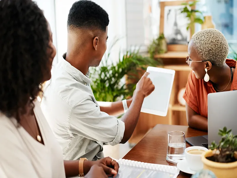 Three people sit at a table with a laptop and documents whilst one person points at a tablet, discussing SMME funding in a bright, modern office. - Proudly Developing
