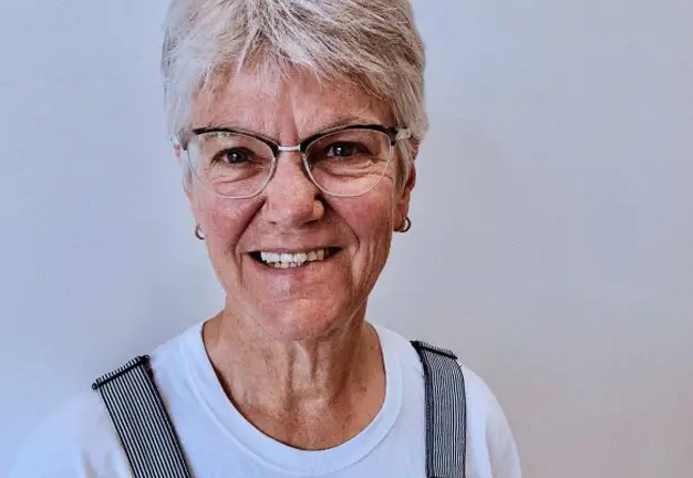 Home 13 Smiling older woman with short grey hair and glasses, wearing a white tee and striped overalls.