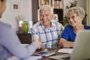 An older man and woman sit at a desk, smiling and shaking hands with another person—reviewing papers, a tablet, and calculator as they discuss using a Reverse Mortgage to Fund Your Retirement.