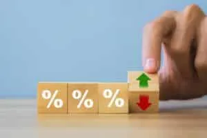 A hand flips a wooden block with a red down arrow and green up arrow, highlighting the effects of interest rates on mortgages, next to three blocks bearing percent signs on a table.