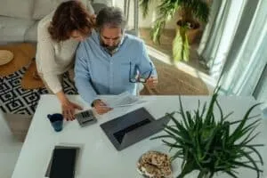 A man and woman review documents at a table with a laptop, calculator, coffee mug, and plate of cookies in a bright, plant-filled room while discussing Conventional Loans for Second Homes.