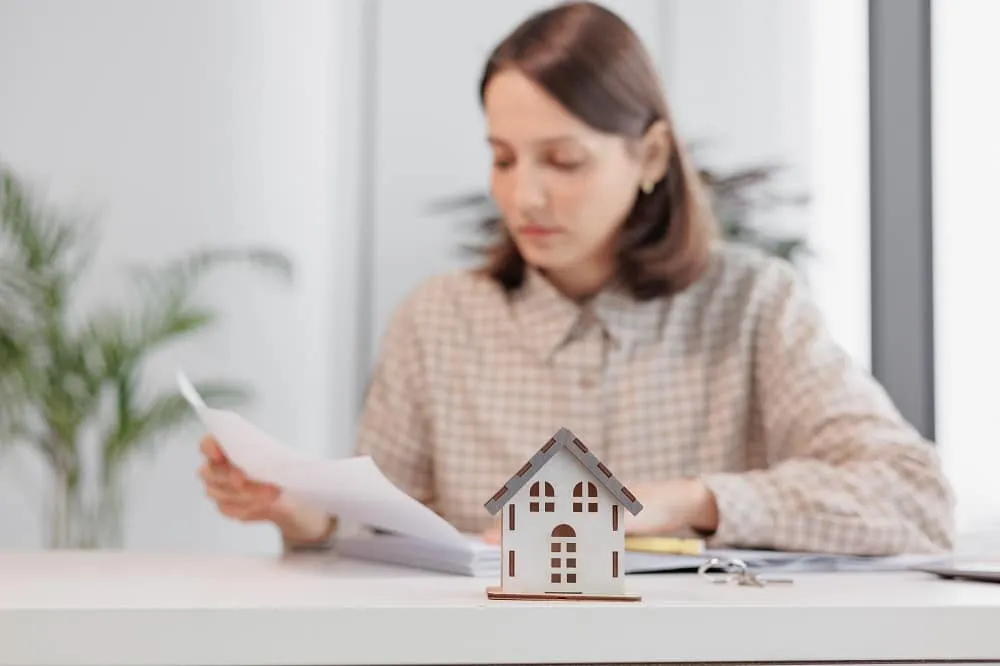 A woman sits at a desk reviewing documents, with a small model house and keys in the foreground—illustrating the careful steps involved in the Bridge Loan Refinancing Timeline.