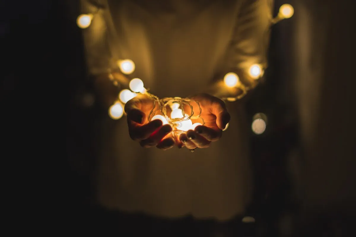 Eine Person hält ein Bündel leuchtender Lichterketten in den Händen, die Hoffnung und Verbundenheit symbolisieren - ein Bild, das an Themen aus der systemischen Therapie anknüpft -, während weitere Lichter vor einem dunklen Hintergrund um ihren Oberkörper drapiert sind.