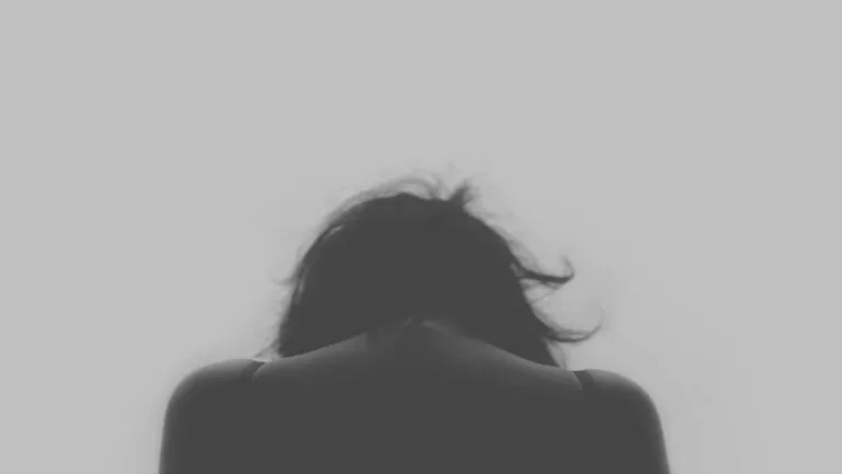 Eine Person mit langen Haaren ist von hinten zu sehen, mit gesenktem Kopf und hängenden Schultern, vor einem schlichten, hellen Hintergrund, der die ruhige Introspektion einfängt, die in der systemischen Therapie oft untersucht wird.