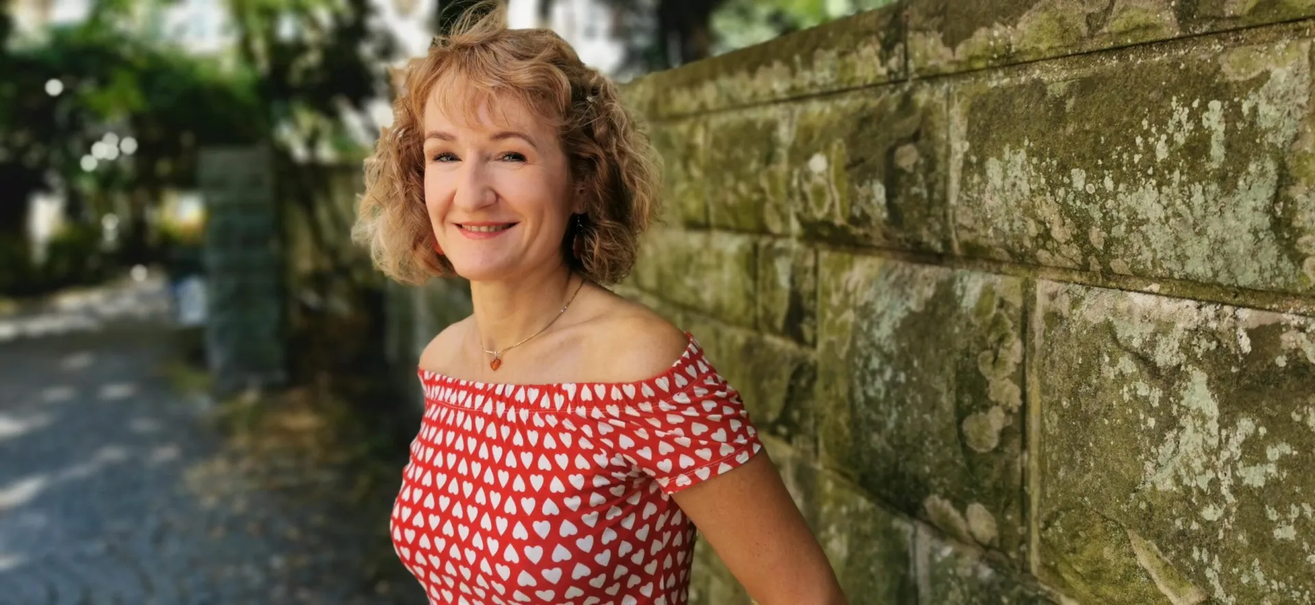 Eine Frau mit lockigem blondem Haar und einem rot-weiß gemusterten Kleid steht lächelnd an einer bemoosten Steinmauer im Freien und strahlt die ruhige Zuversicht aus, die man oft bei denjenigen findet, die systemische Therapie betreiben.