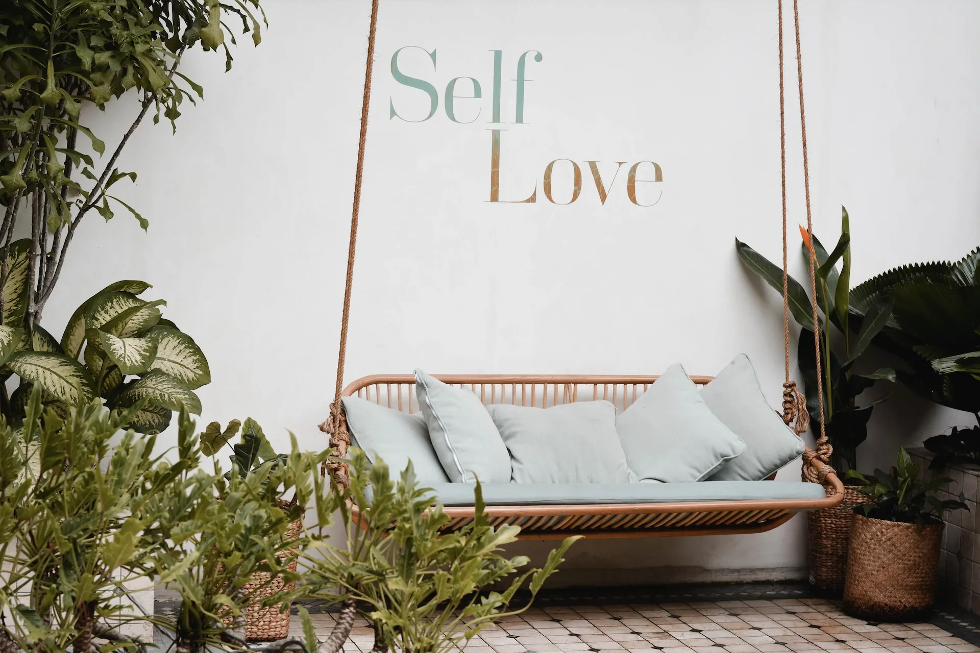 In einem Gartenbereich steht eine Rattan-Schaukelbank mit hellblauen Kissen; auf der weißen Wand dahinter ist der Schriftzug "Self Love" zu lesen, der einen einladenden Raum schafft, der die Prinzipien der systemischen Therapie widerspiegelt.