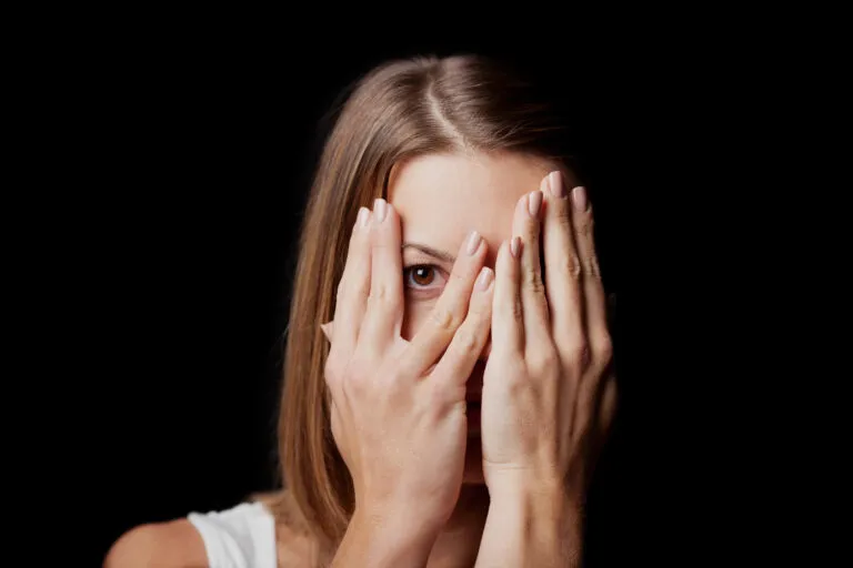 Eine Person mit langem hellbraunem Haar bedeckt ihr Gesicht mit beiden Händen und blickt durch die Finger, als ob sie über Gefühle nachdenkt - ein Konzept, das in der systemischen Therapie erforscht wird. Der Hintergrund ist schwarz.