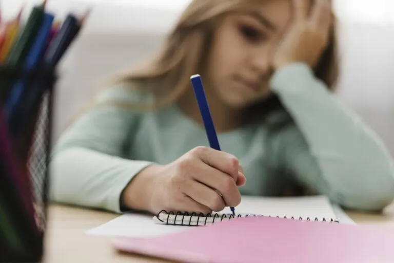 Ein junges Mädchen sitzt an einem Schreibtisch, hält einen blauen Bleistift in der Hand und schreibt in ein Spiralheft, den Kopf auf die Hand gestützt - in Gedanken versunken, vielleicht über Erkenntnisse aus der systemischen Therapie nachdenkend.