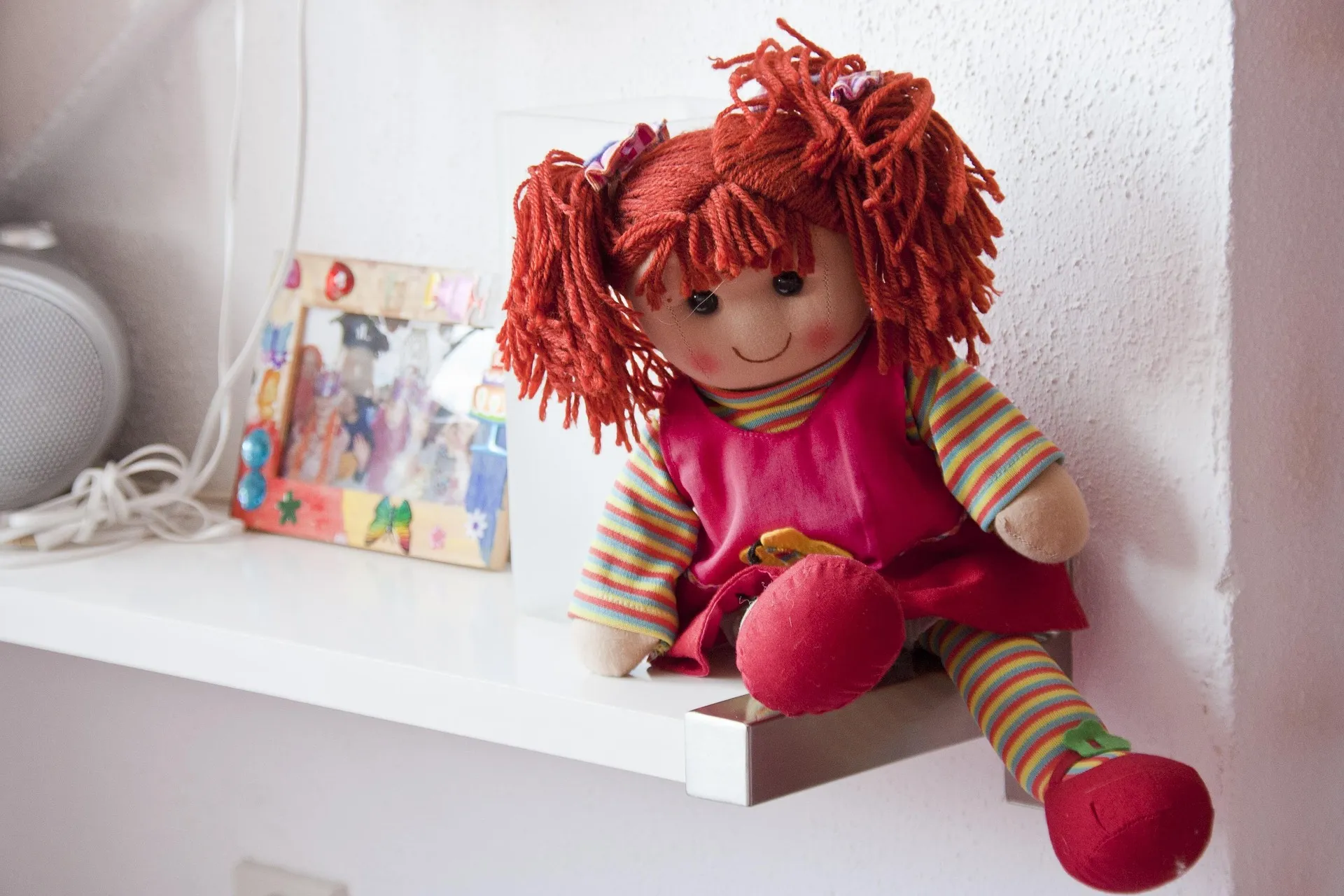 Eine Stoffpuppe mit rotem Garnhaar und einem rosa Kleid steht auf einem weißen Regal neben einem gerahmten Foto und einem Lautsprecher und erinnert an das Spielzeug aus Albert Banduras Bobo-Doll-Experiment.