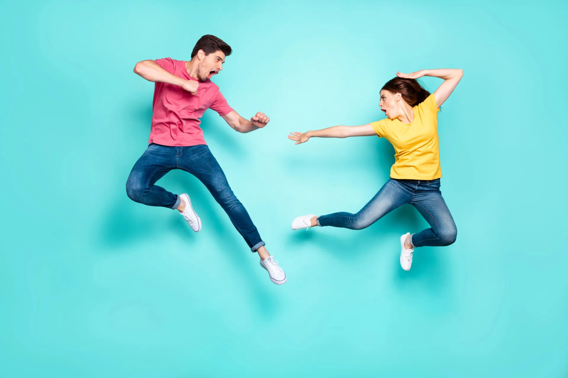 Ein Mann und eine Frau in legerer Kleidung springen in die Luft und stehen sich vor einem türkisfarbenen Hintergrund gegenüber. Ihre dynamischen Posen fangen einen Moment des spielerischen Ärgers oder Grolls ein.