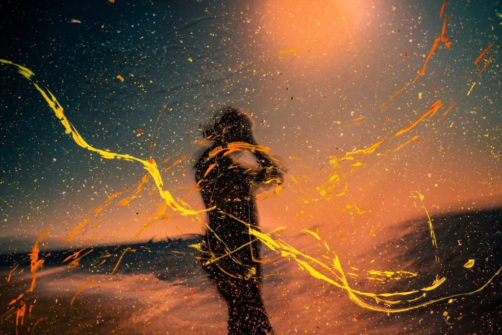 Silhouette einer Person, die bei Sonnenuntergang im Freien steht, überlagert von dynamischen gelben und orangefarbenen Farbspritzern und Schlieren, die die lebendigen Einsichten der systemischen beratung symbolisieren.