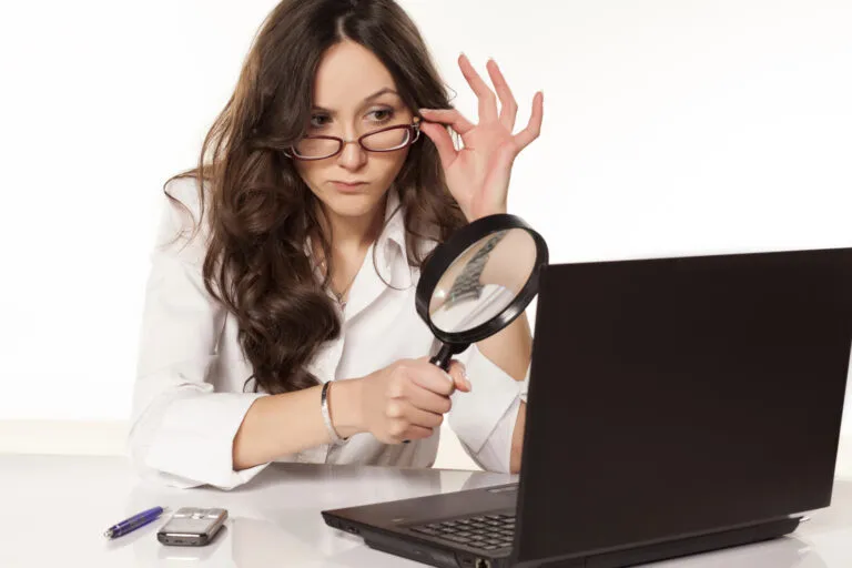 Eine Frau in einem weißen Hemd, die durch eine Lupe auf den Bildschirm eines Laptops schaut, ihre Brille zurechtrückt und auf dem Schreibtisch einen Stift und ein Telefon liegen hat.