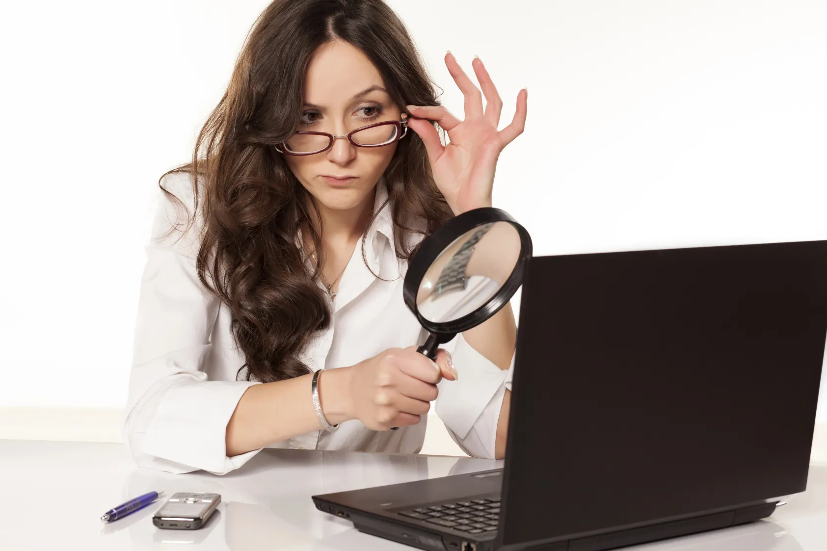 Eine Frau in einem weißen Hemd, die durch eine Lupe auf den Bildschirm eines Laptops schaut, ihre Brille zurechtrückt und auf dem Schreibtisch einen Stift und ein Telefon liegen hat.