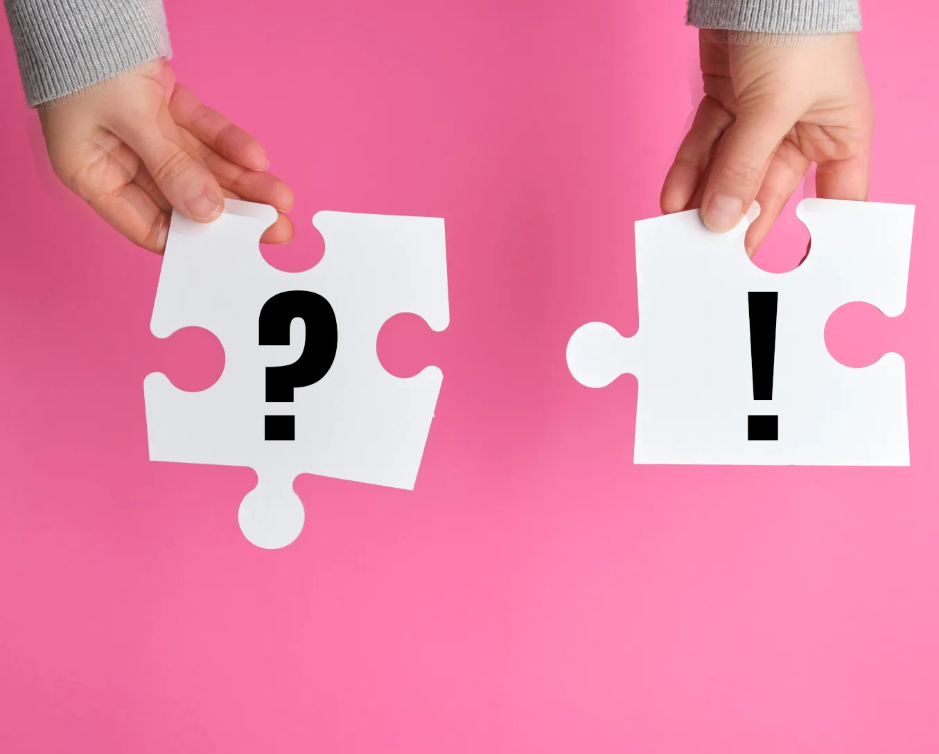 Zwei Hände halten große Puzzleteile vor einem rosa Hintergrund; ein Teil zeigt ein Fragezeichen, das andere ein Ausrufezeichen – als Symbol für den Prozess der Suche nach Antworten und Lösungen bei „Angst Therapie Tübingen“ für Menschen, die mit Ängsten oder Angst- und Panikstörungen zu kämpfen haben, mithilfe systemischer Therapie.