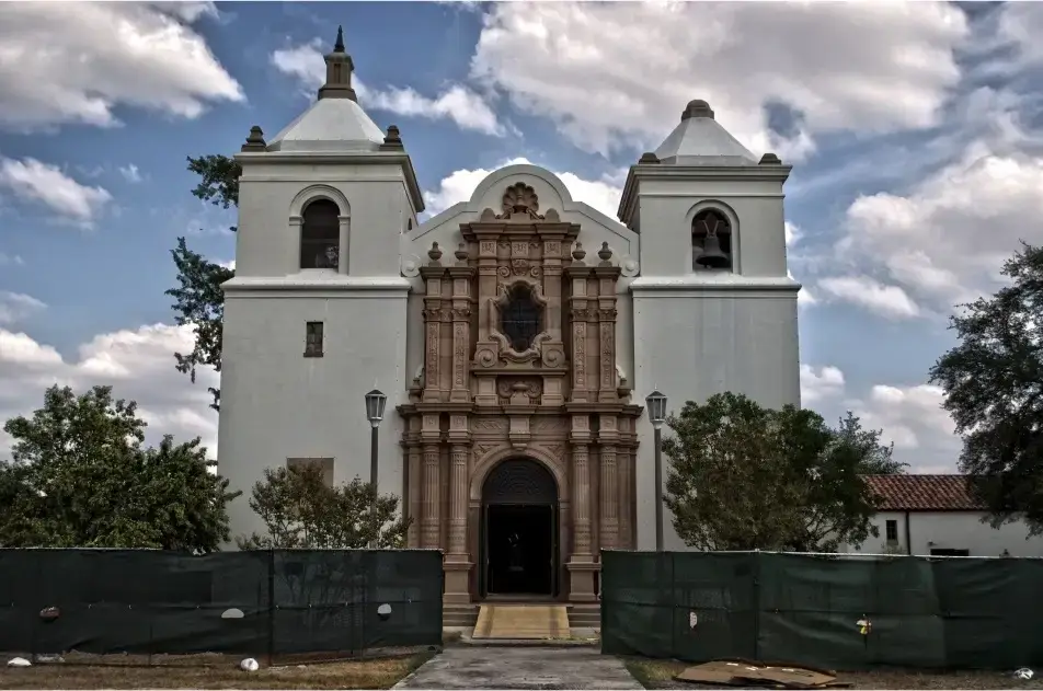 Historic Mission-style building in Universal City, Texas with ornate architectural details, white stucco walls, and bell towers under a partly cloudy sky.