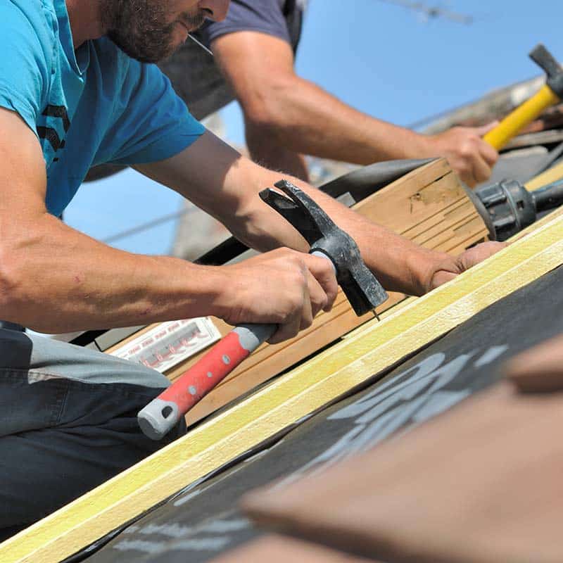 Deux ouvriers du bâtiment utilisent des marteaux pour clouer quelque chose sur une surface en bois à l'extérieur dans le cadre de leurs services de toiture.