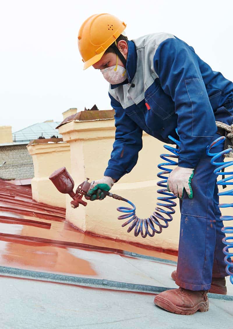 Un travailleur en tenue de protection utilisant un pistolet pulvérisateur pour appliquer de la peinture de toiture sur une toiture métallique.