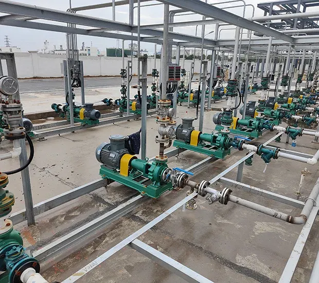 A series of green industrial pumps connected by pipes on a concrete floor, with metal framework above, in an outdoor facility.