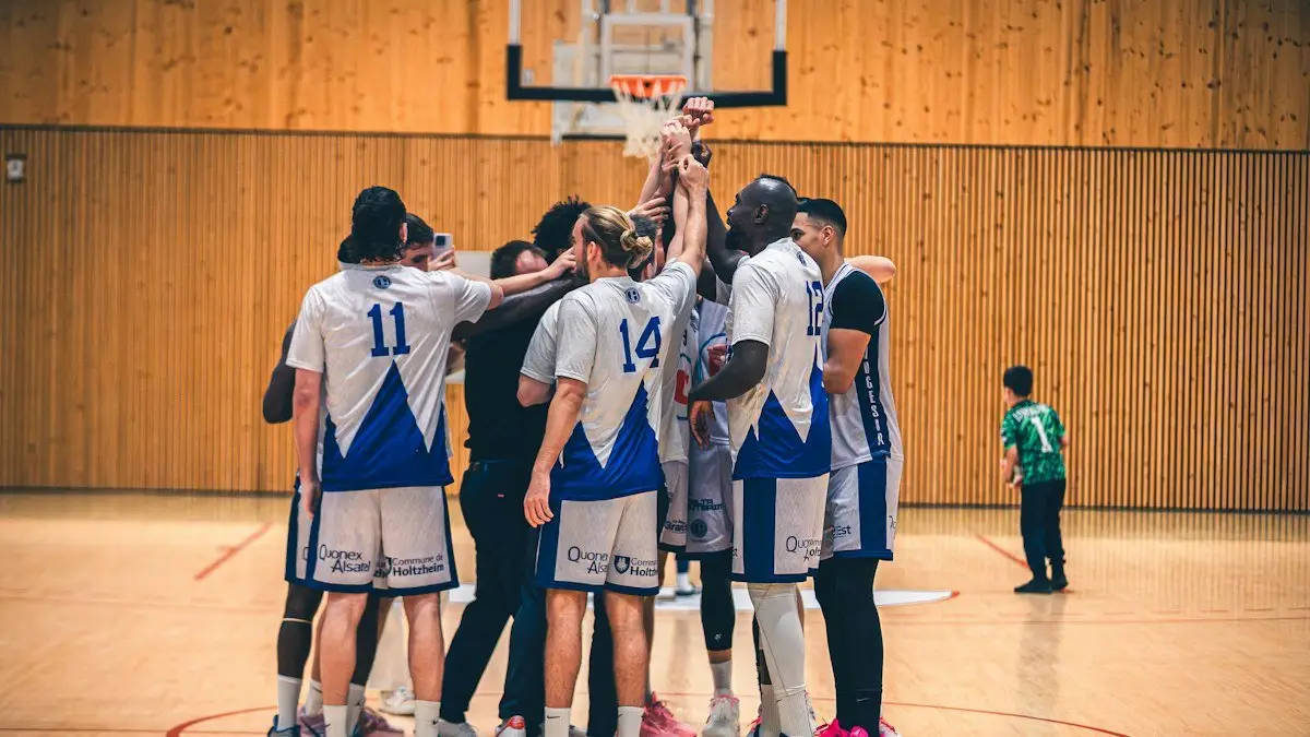 A basketball team in white and blue uniforms gathers in a huddle with hands raised on an indoor court