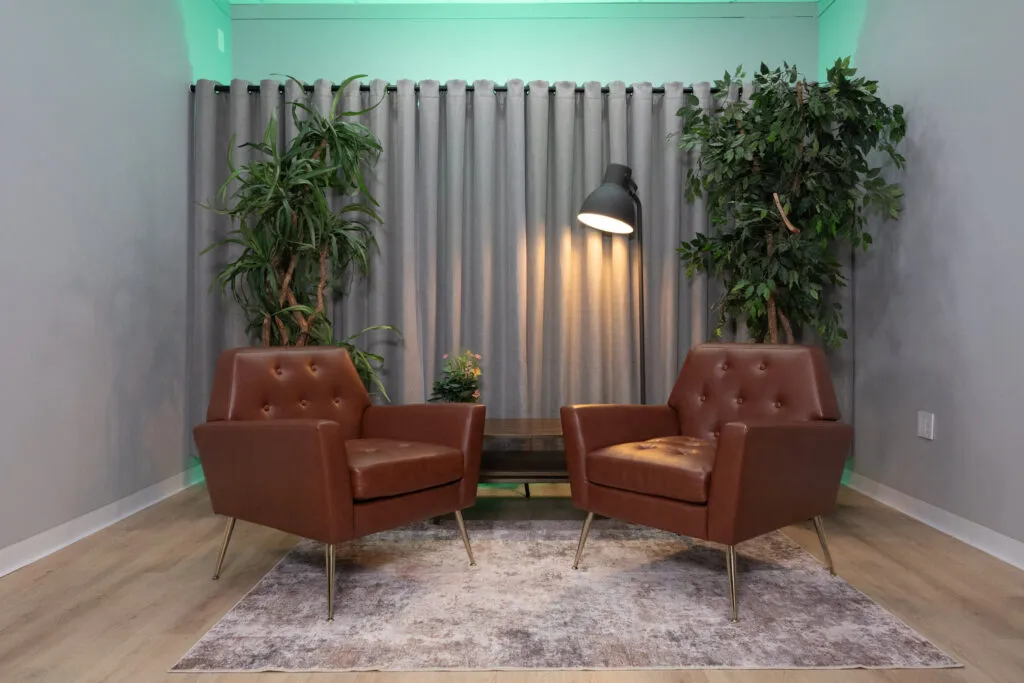 Mid-century leather chairs and indoor plants in a soundproofed Houston podcast and video interview studio with green ambient lighting.