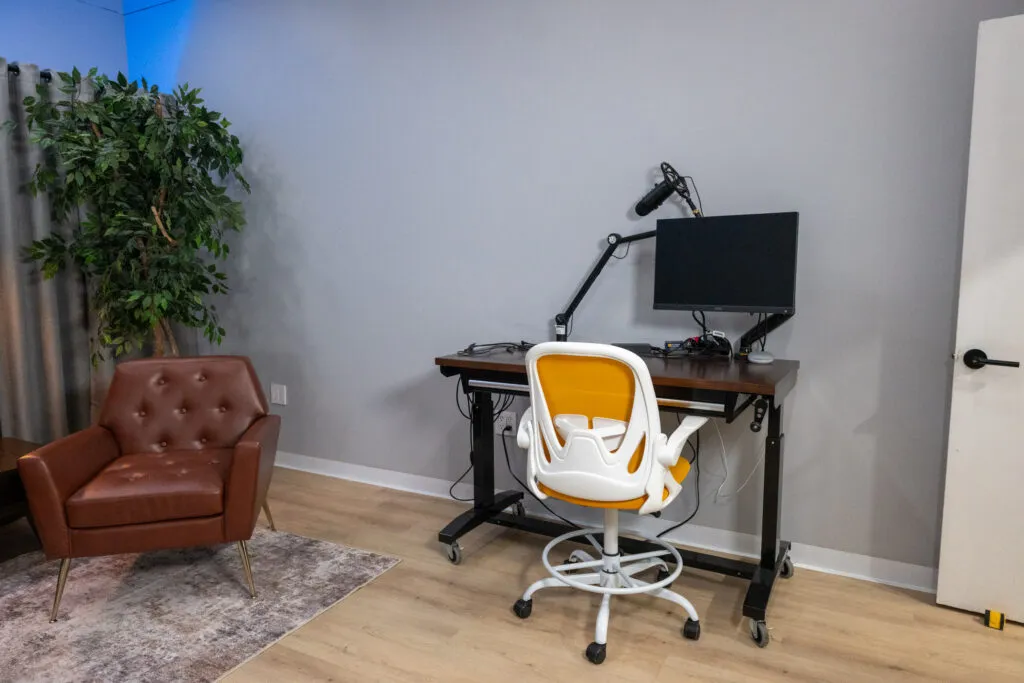 Soundproofed Houston podcast and interview studio with adjustable standing desk, microphone arm, monitor, and orange drafting chair.