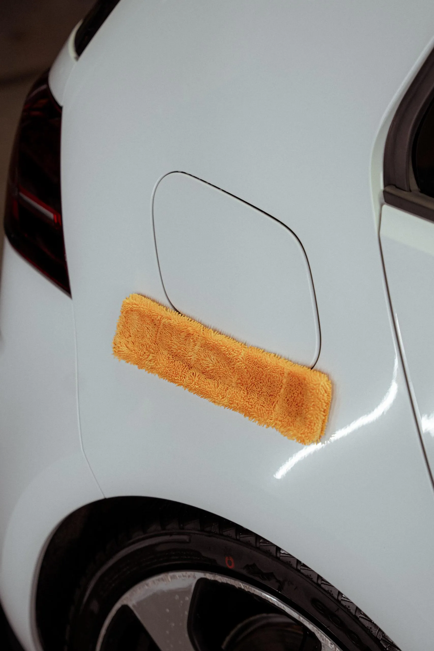A microfiber towel for car detailing is pressed against the rear side below the fuel door of a white car.