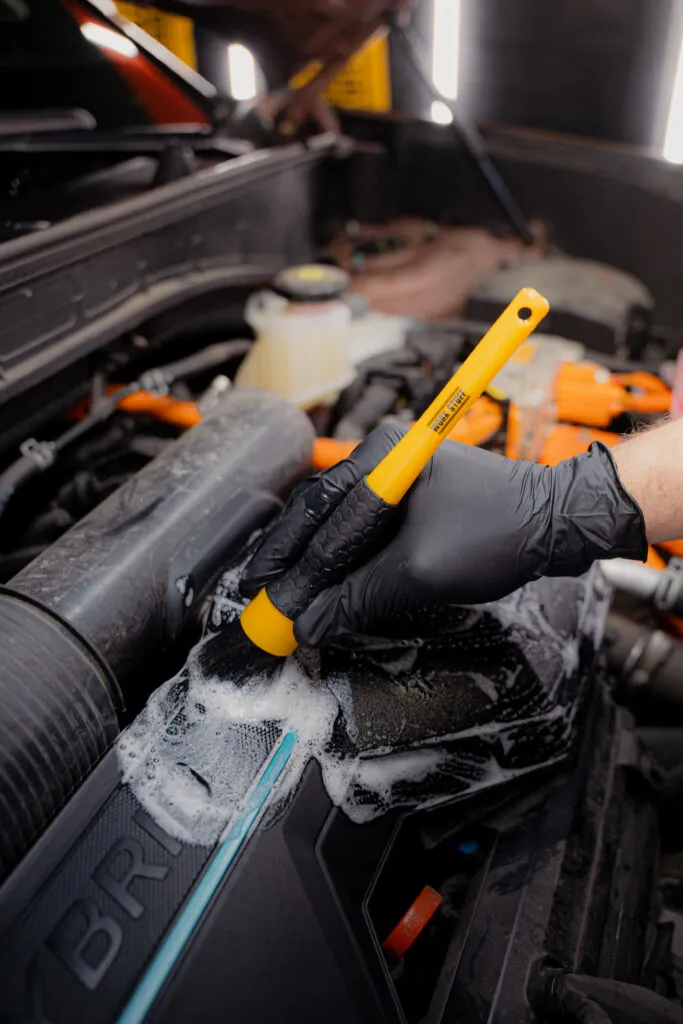 A person in black gloves scrubs a car engine cover with a yellow brush and soapy foam, engine bay open and visible. A person in black gloves scrubs a car engine cover with a yellow brush and soapy foam, engine bay open and visible.