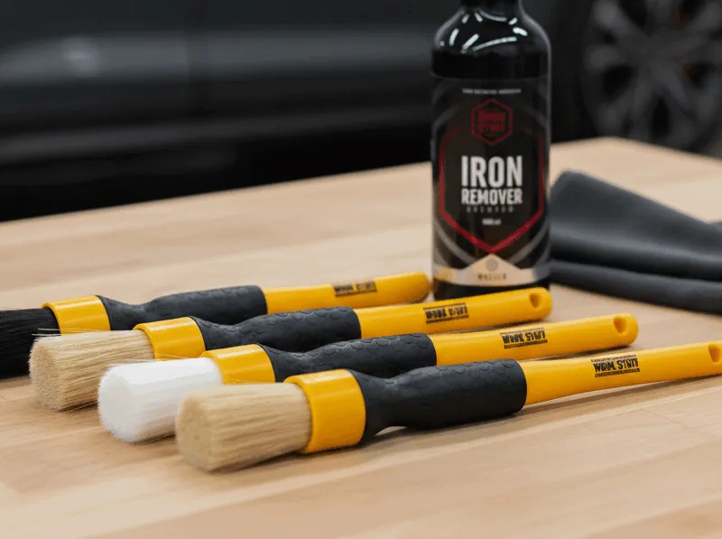 Five yellow-handled detailing brushes, Iron Remover spray, and a folded gray cloth on a wooden surface; car blurred behind.