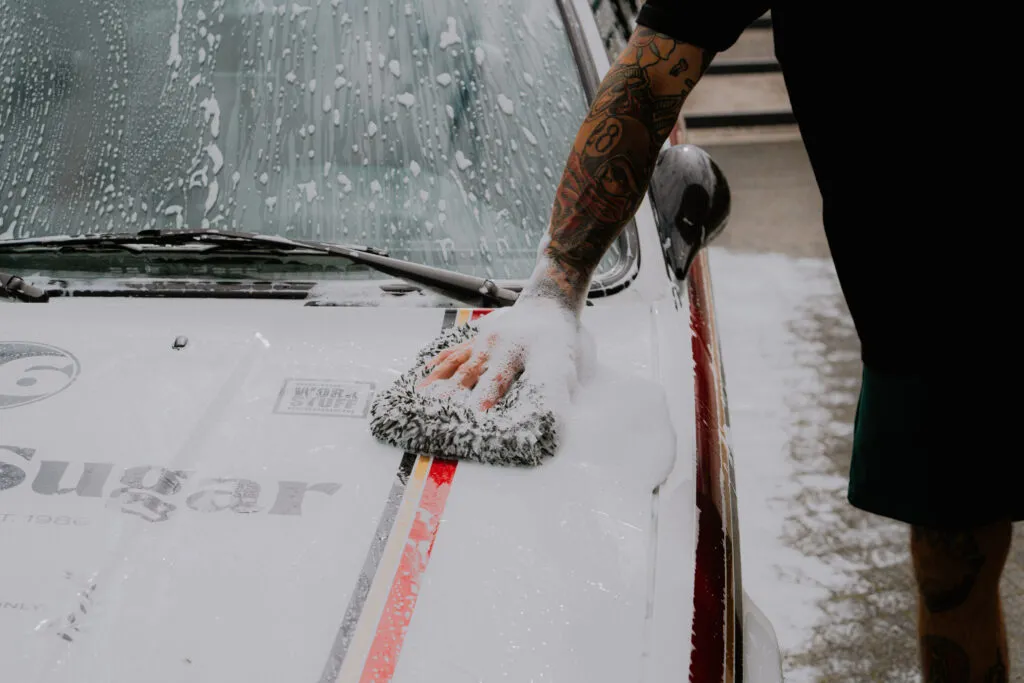 A tattooed person cleans the sudsy hood of a white car with red and black racing stripes using a soapy sponge. A tattooed person cleans the sudsy hood of a white car with red and black racing stripes using a soapy sponge.