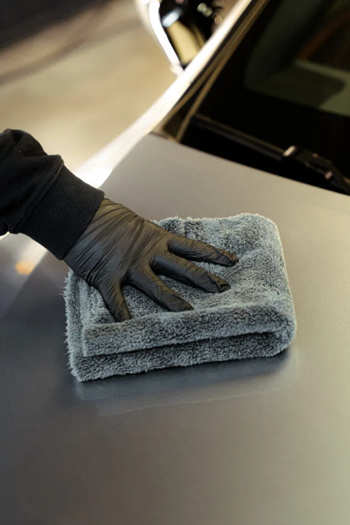 Persona con guante negro limpia la superficie de un coche con un paño gris de microfibra doblado, dejando brillo.