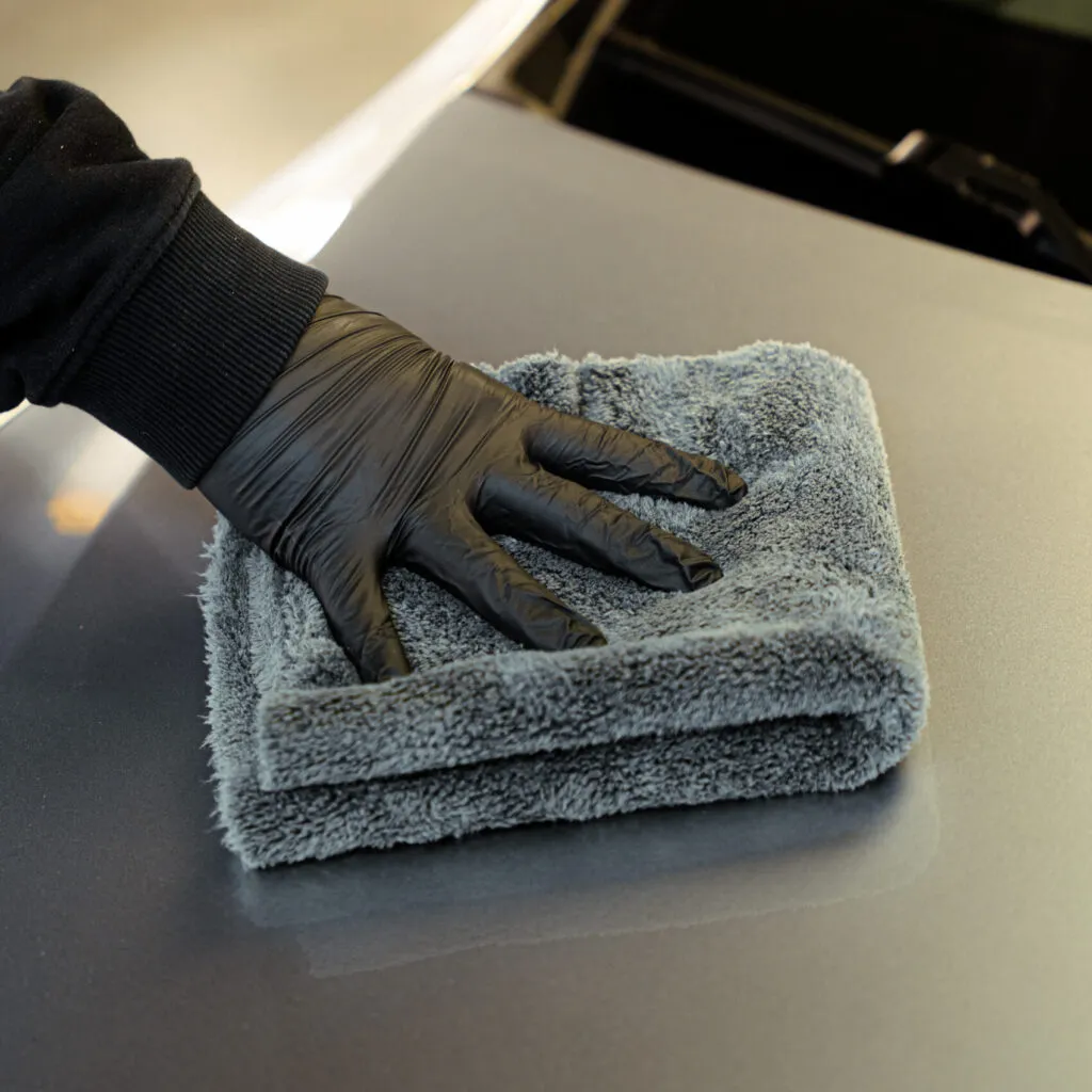 A person in a black glove wipes a car’s surface with a folded gray microfiber cloth, carefully polishing the paint.