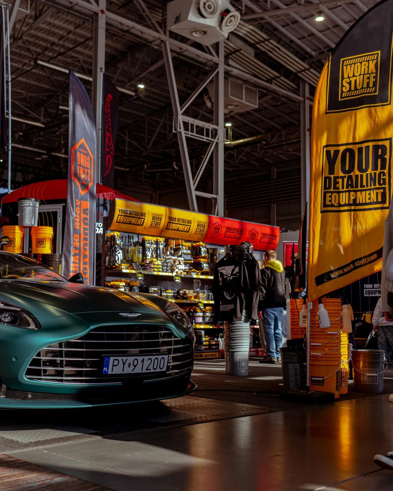 Front of a green car with license plate at AutoCosmetica 2026, yellow orange banners, Work Stuff shelves, people browsing.