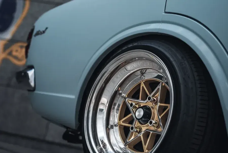 Classic car rear quarter with shiny gold and chrome alloy wheel, low-profile tire; concrete wall with graffiti behind.