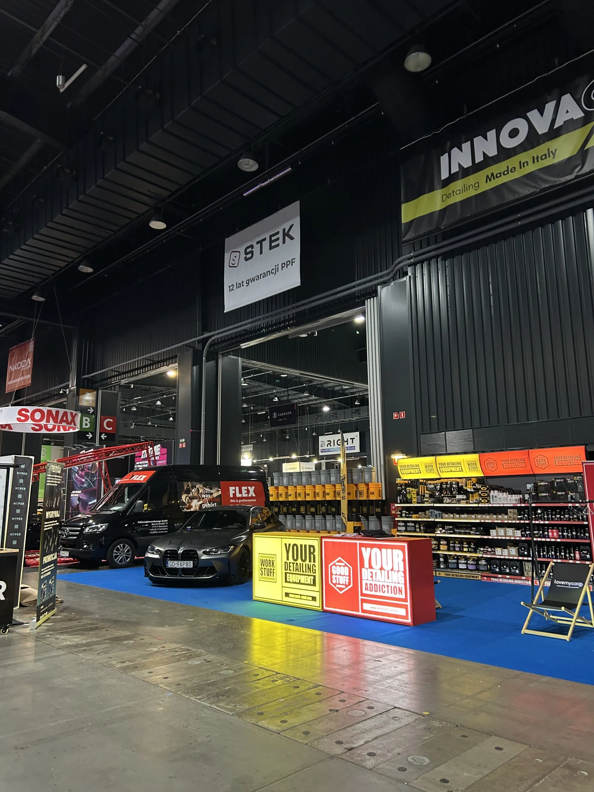A car detailing expo booth with colorful banners, product shelves, and a black car under large Work Stuff signs. Other stalls visible.