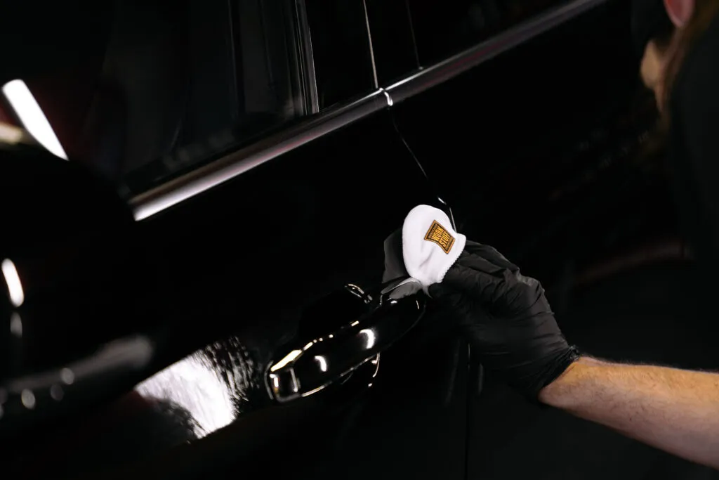 A person wearing a black glove uses a white cloth to clean or polish the handle of a black car.