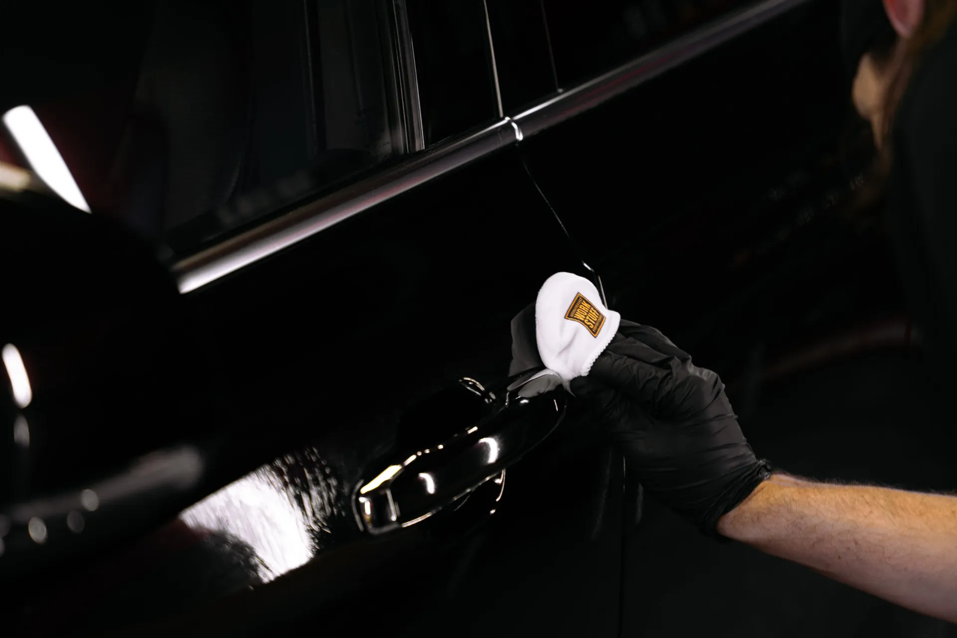 A person wearing a black glove uses a white cloth to clean or polish the handle of a black car.