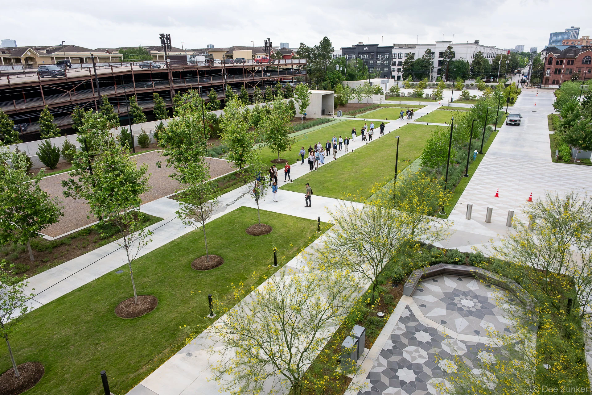 Elevated view of The Ismaili Center Houston grounds along Allen Parkway, with conference attendees walking geometric pathways amid green lawns and young trees.