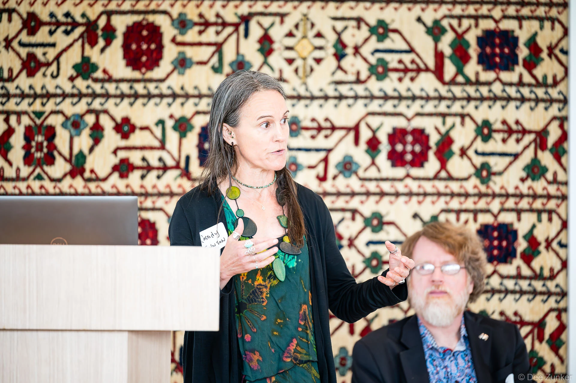 Dr. Cassidy Johnson speaker gestures at a podium during a panel discussion at Gulf Coast Green 2026, Ismaili Center Houston, ornate tapestry behind her.