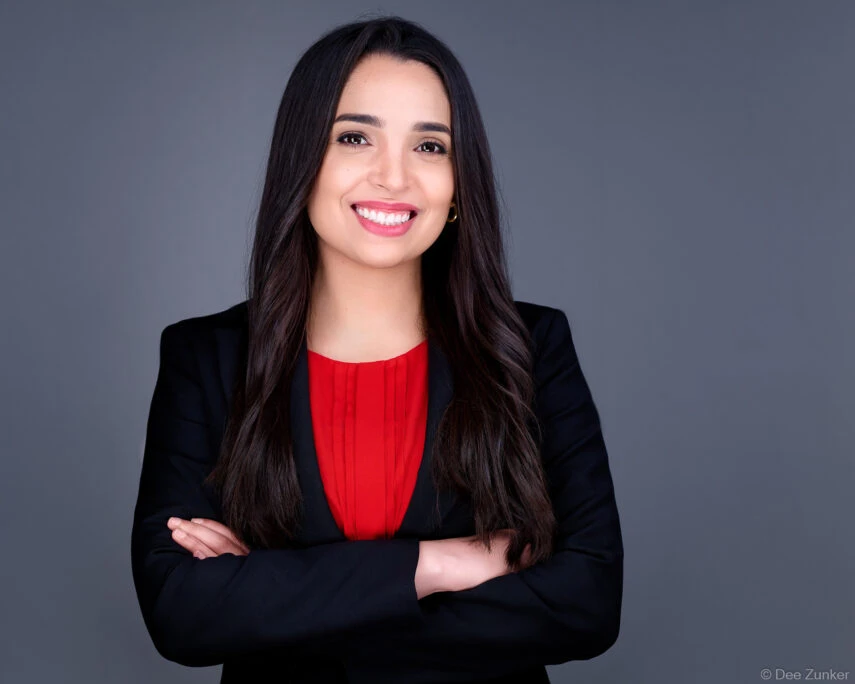 Female attorney in black blazer and red top poses with arms crossed for a professional studio headshot in Houston, gray background.