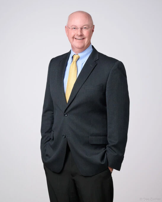 Professional corporate headshot of a smiling male financial advisor in a dark suit and gold tie, studio portrait with gray background, Houston TX.