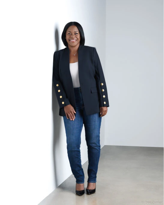 Full-length corporate headshot of a smiling professional woman in a navy blazer and jeans, leaning against a white studio wall in Houston.
